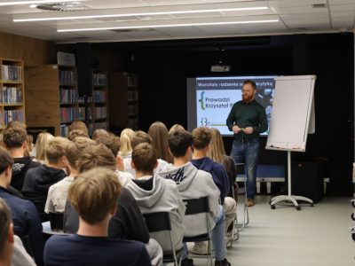 Krzysztof Piersa stojący obok czystej tablicy flipchart przemawia do grupy młodzieży zebranej w bibliotece. Za nim wyświetla się slajd tytułowy prezentacji