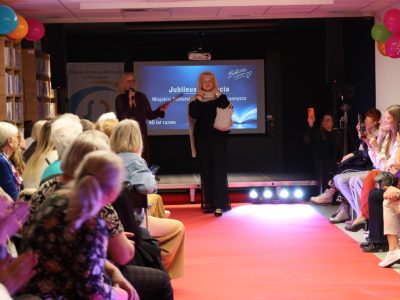 Uśmiechnięta modelka w czarnym komplecie stoi na wybiegu i prezentuje jasną, dzianinową torebkę, trzymając ją przed sobą obiema rękami. Zgromadzona widownia bije brawo