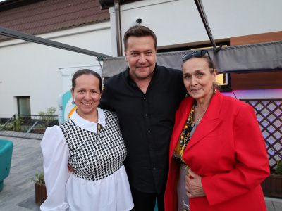 Pamiątkowa fotografia Roberta Kudelskiego stojącego pomiędzy dwiema kobietami. Po jego prawej stronie stoi kobieta w białej bluzce z wzorem w czarno-białą jodełkę, a po lewej kobieta w wyrazistej, czerwonej marynarce z okularami przeciwsłonecznymi na głowie. Wszyscy troje uśmiechają się szeroko do aparatu na tle plenerowej sceny