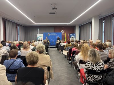 Szerokie ujęcie z perspektywy tyłu sali na licznie przybyłych uczestników spotkania, przysłuchujących się gościom na scenie pod niebieską ścianką Powiatu Przasnyskiego