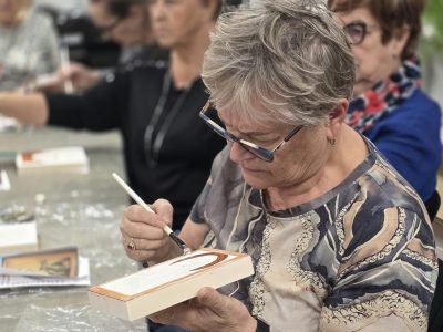Kobieta w okularach i wzorzystej bluzce trzyma w dłoni drewnianą deseczkę i precyzyjnie maluje pędzelkiem jej krawędzie