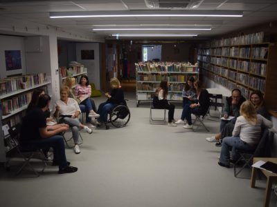 Widok na całą salę biblioteczną z regałami po bokach. Kilka małych grup uczestników szkolenia siedzi na krzesłach w różnych częściach pomieszczenia i pracuje nad zadaniami w podgrupach