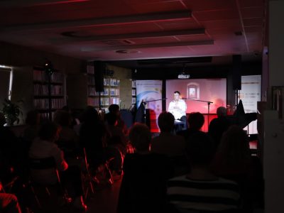Szerokie ujęcie przyciemnionej sali widowiskowej biblioteki podczas występu. Na oświetlonej czerwonym światłem scenie występuje mężczyzna w białej koszuli. Wypełniona po brzegi sala jest widoczna z perspektywy tyłu – z wyraźnymi zarysami sylwetek siedzących widzów