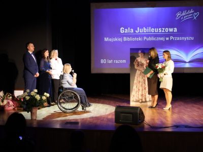 Kobieta w ciemnej bluzce przemawia do mikrofonu trzymając zieloną teczkę z dyplomem, a stojąca obok niej kobieta w jasnym komplecie trzyma bukiet kwiatów skierowany w stronę dyrektor