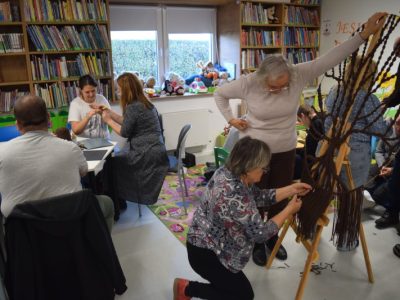 Uczestnicy zajęć pracują w grupach w przestrzeni bibliotecznej. Dwie kobiety przycinają i wykańczają dolne frędzle brązowego drzewa z makramy opartego na sztaludze. Przy stolikach w głębi sali inne osoby siedzą tyłem i bokiem, zajmując się drobnymi pracami manualnymi przy użyciu kolorowych kartek