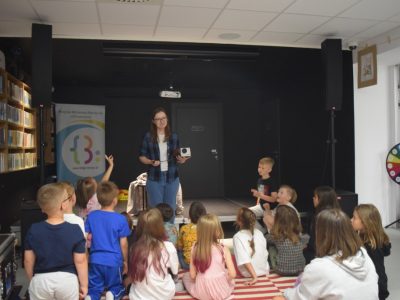 Kobieta w rozpiętej koszuli w kratę stoi na niskim podwyższeniu w sali bibliotecznej i trzyma retro projektor, przemawiając do grupy dzieci siedzących przed nią tyłem na pasiastym dywanie