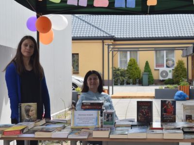 Dwie kobiety stoją za stołem charytatywnego kiermaszu książek pod zadaszeniem. Stół jest gęsto zastawiony różnorodnymi książkami wystawionymi na sprzedaż. Na środku stołu stoi zalaminowana kartka z informacją: „Kiermasz książek. Całkowity dochód z kiermaszu zostanie przeznaczony na leczenie Krzysztofa Nowakowskiego”. W tle widoczny jest jasny budynek oraz kostka brukowa na dziedzińcu