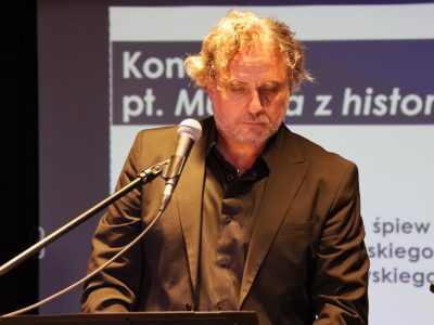 Bliskie ujęcie mężczyzny z kręconymi włosami, w ciemnym garniturze i czarnej koszuli, stojącego przy mikrofonie ze spuszczonym wzrokiem. W tle widać fragment slajdu z tekstem „Koncert pt. Muzyka z historią w tle”