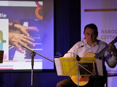 Mężczyzna w jasnej koszuli gra na gitarze klasycznej, siedząc przed pulpitem z nutami. W tle widoczny jest ekran projekcyjny ze slajdem prezentacji oraz fragment napisu „Narodowe Czytanie”