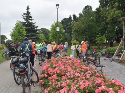 Grupa uczestników rajdu rowerowego z rowerami stoi na placu miejskim za pasem bujnie kwitnących, różowych róż. Część osób ma na sobie pomarańczowe i żółte kamizelki odblaskowe. W tle widoczne są zielone drzewa, ławki parkowe oraz wysoka latarnia