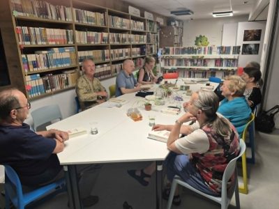 Uczestnicy spotkania czytelniczego siedzą przy podłużnym, białym stole w bibliotece. Zdjęcie wykonano z perspektywy ukazującej tył głowy jednego z mężczyzn na pierwszym planie. Pozostałe osoby słuchają wypowiedzi lub przyglądają się książkom leżącym na stole. W tle widać regały z literaturą oraz rysunek zielonego smoka na dalekiej ścianie