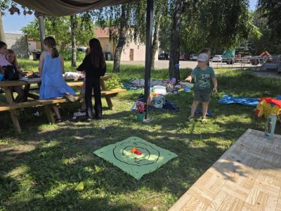 Grupa dzieci spędza czas w plenerze obok zadaszonej altany. Część dzieci siedzi przy drewnianym stole biesiadnym, podczas gdy chłopiec w zielonej koszulce i czapce z daszkiem rzuca kolorowym woreczkiem do celu – zielonej maty z narysowanymi kręgami, leżącej na trawie. W tle widoczne są drzewa i budynki