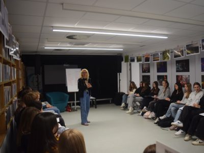 Joanna Jagiełło prowadzi warsztaty dla młodzieży w bibliotece, stojąc w centrum utworzonego przez uczestników kręgu. Dziewczęta i chłopcy siedzą na krzesłach pod ścianami, na których wiszą wystawy zdjęć. Wszyscy są skupieni na słowach prowadzącej, niektórzy trzymają na kolanach notatniki i karty pracy
