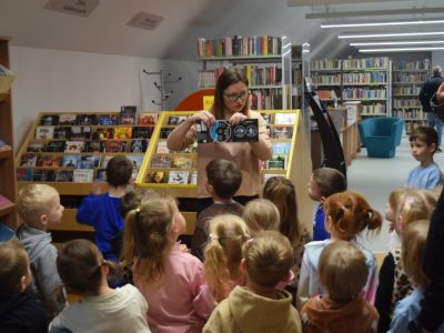 Bibliotekarka w okularach stoi przed grupą małych dzieci w dziale zbiorów audiowizualnych. Kobieta pokazuje dzieciom otwarte opakowanie z płytą CD, tłumacząc zasady wypożyczania multimediów. Dzieci z zaciekawieniem patrzą na prezentację