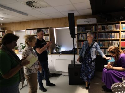 Maria Paszyńska podpisuje książkę przy stoliku w bibliotece. Obok niej stoi kobieta w jeansowej kurtce, a po lewej stronie w kolejce czekają inni uczestnicy spotkania, w tym mężczyzna robiący zdjęcie smartfonem. W tle regały pełne książek