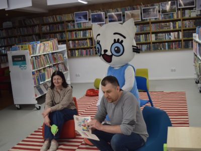 Mężczyzna siedzi na niskim, niebieskim fotele i czyta na głos otwartą książkę z obrazkami. Za jego plecami stoi maskotka Kici Koci, a po lewej stronie, na czerwonej pufie, siedzi uśmiechnięta kobieta. Scena odbywa się w kąciku dziecięcym w bibliotece, na tle regałów z książkami i pasiastego dywanu