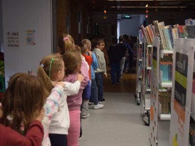 Dzieci idą jedno za drugim w wąskim przejściu między regałami bibliotecznymi, trzymając ręce na ramionach kolegów i koleżanek przed nimi. Na ścianie po lewej stronie wisi plakat z napisem „Fajne dzieciaki czytają książki”