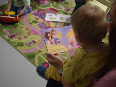 Widok zza ramienia małego dziecka siedzącego na kolanach dorosłego i oglądającego otwartą, kolorową książeczkę obrazkową przedstawiającą ilustrację z radosną rodziną