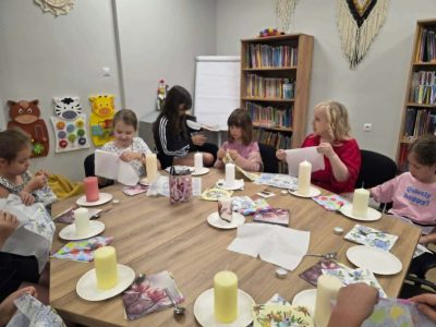 Dzieci oraz jasnowłosa instruktorka w czerwonej bluzce siedzą wokół dużego, wielokątnego stołu i przygotowują papierowe serwetki do zdobienia. Na stole, przed każdym z uczestników, stoi na białym talerzyku duża, jasna świeca walcowata
