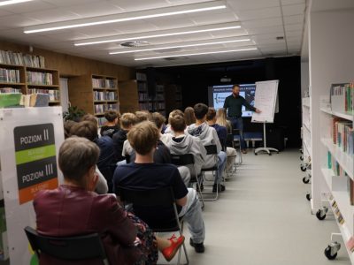 Mężczyzna z brodą w ciemnej koszuli stoi przed młodzieżą w bibliotece i wskazuje na schemat narysowany na flipcharcie. W tle widać ekran z prezentacją „Warsztaty radzenia sobie z krytyką”