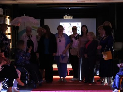 Finał pokazu mody w bibliotece. Grupa kilkunastu kobiet uczestniczących w pokazie stoi razem na scenie na końcu czerwonego dywanu. Jedna z kobiet siedzi na wózku inwalidzkim i mówi do mikrofonu. W tle widoczny jest ekran projekcyjny ze szkicami ubrań