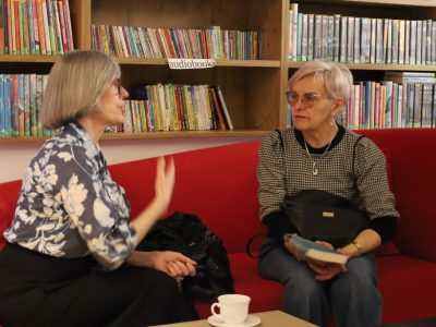 Grażyna Mączkowska siedzi na czerwonej sofie w bibliotece, gestykulując podczas rozmowy z drugą kobietą. W tle znajdują się regały z książkami