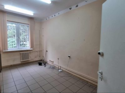 Wnętrze pustego pokoju biurowego lub magazynowego, widoczne z perspektywy uchylonych białych drzwi. Pomieszczenie ma podłogę z szarych płytek i jasne ściany. Wzdłuż prawej ściany, tuż przy podłodze, ciągną się kable i leży biały router. Na ścianie pod sufitem widoczne są ślady po wierceniu lub ubytki w tynku. W głębi pokoju znajduje się okno
