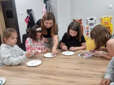 Grupa dzieci siedzi przy drewnianym stole i w skupieniu nawleka kolorowe koraliki z talerzyków na sznurki. Kobieta w okularach pochyla się nad nimi, pomagając jednej z dziewczynek