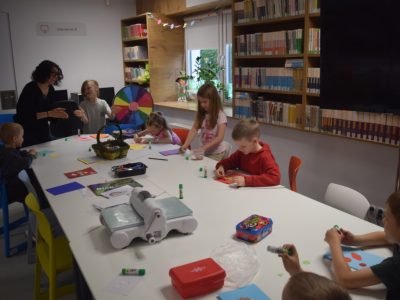 Grupa dzieci siedzi przy dużym białym stole w bibliotece i wykonuje kartki okolicznościowe metodą scrapbookingu. Na stole leżą kolorowe papiery, kleje w sztyfcie i maszynka do wycinania wzorów. W tle widać regały z książkami oraz uśmiechniętą instruktorkę rozmawiającą z dziećmi