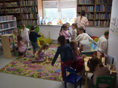 Wnętrze biblioteki dla dzieci. Na pierwszym planie dzieci siedzą przy małym stoliku i rysują. W tle dzieci bawią się na kolorowym dywanie z nadrukiem mapy miasta, przy regałach wypełnionych książkami. Kobieta stoi przy regale, pomagając dziecku w wyborze książki