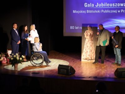 Mężczyzna w szarej marynarce przemawia do mikrofonu, trzymając zieloną teczkę, a stojący obok niego mężczyzna trzyma pamiątkowy dyplom. Zwracają się oni w stronę dyrektor biblioteki siedzącej na wózku inwalidzkim w otoczeniu prowadzących wydarzenie. W tle widać portret historyczny oraz ekran z napisem „Gala Jubileuszowa Miejskiej Biblioteki Publicznej w Przasnyszu”