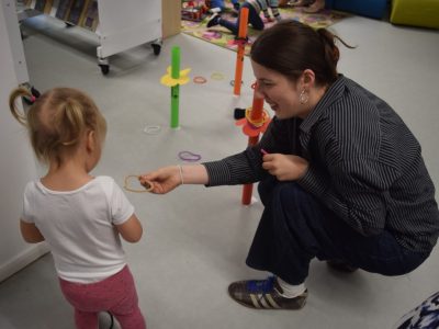 Wolontariuszka w koszuli w paski kuca na dywanie i podaje kolorową obręcz małej dziewczynce spiętej w dwa kucyki. Wokół na podłodze rozstawione są plastikowe stojaki do gry zręcznościowej