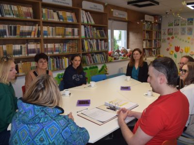 Kadrowane z bliska ujęcie uczestników spotkania siedzących przy stole w bibliotece. Na pierwszym planie widoczny jest mężczyzna w czerwonej koszulce oraz kobieta w niebiesko-zielonej wzorzystej bluzie. W tle znajdują się wysokie drewniane regały ścienne z książkami oraz okno, na którego parapecie stoją pluszowe maskotki