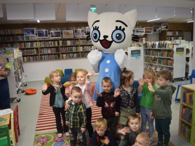 Grupa uśmiechniętych dzieci pozuje do zdjęcia wspólnie z dużą maskotką Kici Koci w jasnoniebieskiej sukience. Niektóre dzieci machają rękami w stronę obiektywu. Po lewej stronie stoi uśmiechnięta kobieta w pasiastej koszuli, trzymająca książkę. W tle widoczne są regały z książkami oraz galeria zdjęć na ścianie