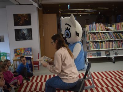 Kobieta siedzi na krześle tyłem do kadru i czyta książkę grupie przedszkolaków siedzących na dywanie w bibliotece. Obok niej, na drugim krześle, siedzi postać w dużym kostiumie maskotki Kici Koci, która patrzy w stronę dzieci. W tle widać białe regały wypełnione literaturą dziecięcą