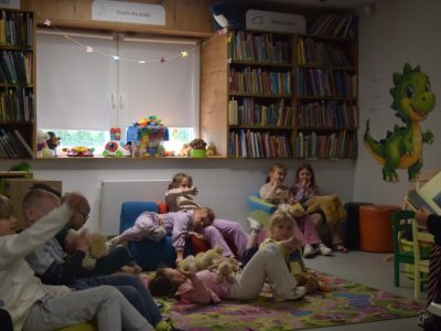 Widok na salę biblioteczną z perspektywy strefy dla dzieci. Sylwia Błach, czyta książkę grupie kilkorga dzieci siedzących na dywanie i kolorowych siedziskach. Na ścianach wiszą plakaty, a na półkach pod oknem leżą zabawki i klocki. Panuje przytulna, skupiona atmosfera warsztatów literackich