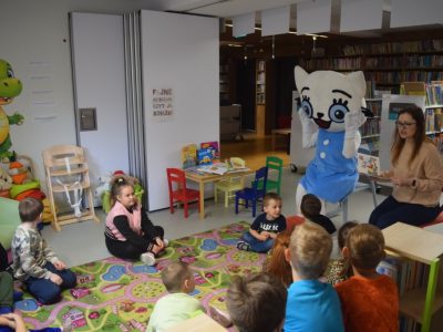 Grupa dzieci siedzi na kolorowym dywanie w kąciku dziecięcym i słucha bajki. Bibliotekarka siedzi obok maskotki Kici Koci i czyta na głos książeczkę. Maskotka Kicia Kocia przykłada ręce do policzków w geście zdziwienia, animując czytaną historię