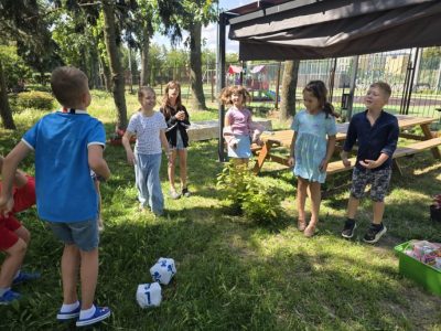 Radosna grupa dzieci bawi się na trawniku w ogrodzie pod ciemnym zadaszeniem altany. Na trawie w pobliżu chłopca w niebieskiej koszulce leżą dwie duże, białe, pluszowe kostki do gry, a w tle widoczny jest ogrodzony plac zabaw