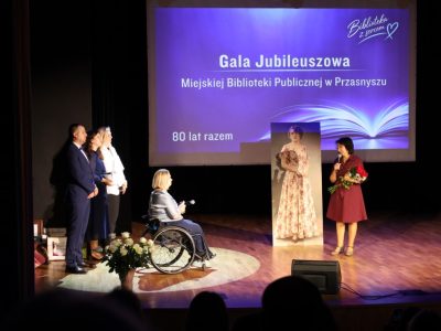 Kobieta w eleganckiej bordowej sukience przemawia do mikrofonu, trzymając w drugiej dłoni bukiet czerwonych róż zwrócony w stronę dyrektor biblioteki