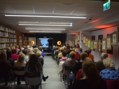 Perspektywiczny widok na długą salę Miejskiej Biblioteki Publicznej w Przasnyszu wypełnioną publicznością. Po lewej stronie znajdują się regały z książkami, po prawej galeria rysunków, a na końcu sali, na podwyższeniu, trwa spotkanie z Arturem Dziurmanem