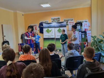 Ujęcie zza pleców widowni na dwóch chłopców stojących na środku sali obok prowadzących w koszulkach piłkarskich. W tle wiszą koszulki znanych piłkarzy oraz baner rolowany z napisem „Ekstraklasa czyta”
