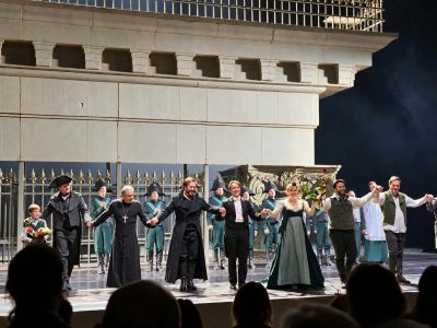 Aktorzy i śpiewacy w strojach epokowych trzymają się za ręce i ukłonią widowni na scenie teatru na tle monumentalnej dekoracji. Jedna z aktorek trzyma czerwoną różę, a chłopiec bukiet kwiatów