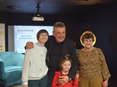 Aktor Artur Dziurman pozuje do pamiątkowego zdjęcia z trzema uczestniczkami spotkania – dwiema kobietami i uśmiechniętą dziewczynką w czerwonej bluzce, którą mężczyzna obejmuje. W tle widoczny ekran z napisem „Spotkanie z Arturem Dziurmanem”