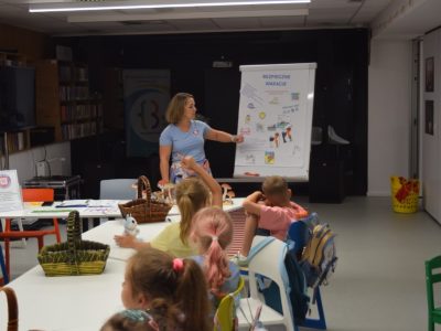Kobieta stoi przy tablicy flipchart z napisem „Bezpieczne wakacje” i wskazuje na przypięte do niej rysunki edukacyjne. Przed nią, przy białym stole, siedzi tyłem grupa dzieci