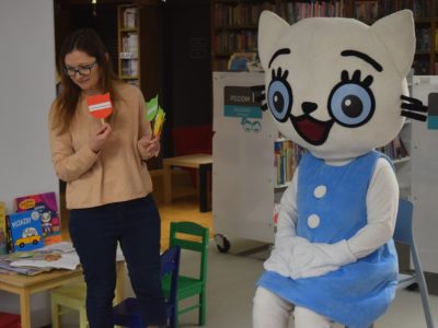 Maskotka Kici Koci siedzi na wysokim krześle w bibliotece. Obok niej stoi bibliotekarka, która trzyma w rękach kolorowe rekwizyty w kształcie głowy kota na patyczkach. Na małym stoliku obok leżą książeczki z serii o Kici Koci