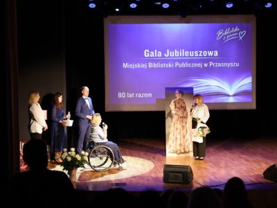 Widok na scenę z perspektywy widowni, na której kobieta w bluzce w groszki z bukietem kwiatów przemawia do dyrektor biblioteki na wózku inwalidzkim
