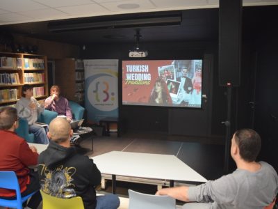 Spotkanie kawiarenki językowej w bibliotece. Dwie prowadzące siedzą na fotelach i przemawiają do słuchaczy, a na ekranie projektora wyświetla się slajd z napisem „Turkish Wedding Traditions”