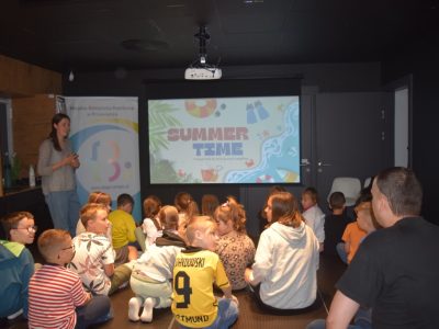 Prowadząca zajęcia stoi przy ekranie projekcyjnym wyświetlającym slajd z napisem „SUMMER TIME”. Na podłodze siedzi grupa dzieci – jedno z nich ma na sobie żółtą koszulkę piłkarską z numerem 9 i nazwiskiem Lewandowski