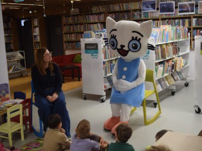 Bibliotekarka siedzi na niebieskim krześle obok siedzącej maskotki Kici Koci. Oboje zwróceni są w stronę grupy dzieci, których fragmenty postaci widać na dole zdjęcia. W tle znajdują się białe, mobilne regały biblioteczne oznaczone jako „Poziom I” i „Poziom II”