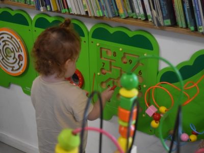 Dziewczynka stoi przy zielonej tablicy edukacyjnej zamontowanej pod półką z książkami i przesuwa elementy po labiryncie z numerami